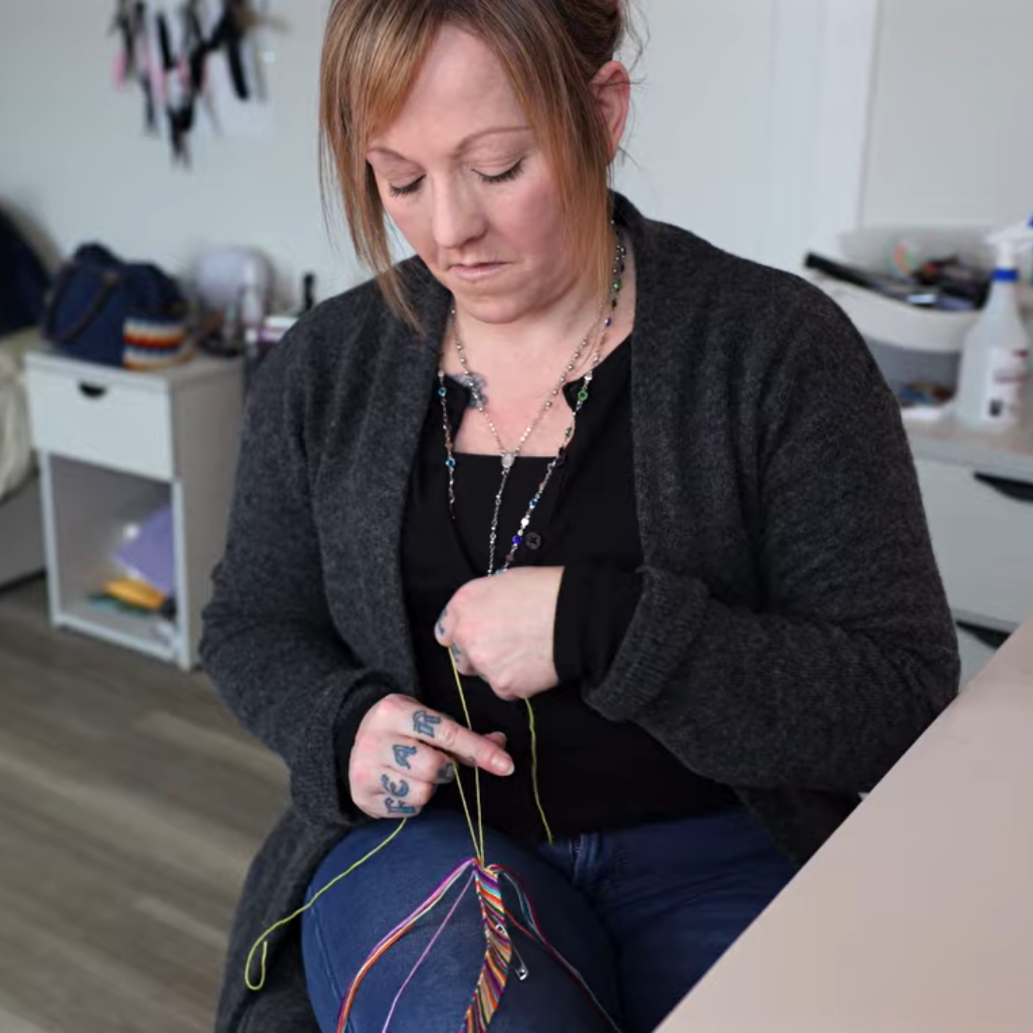A person with blonde hair sitting on a chair indoors by a counter in the kitchen making a multicoloured string bracelet.