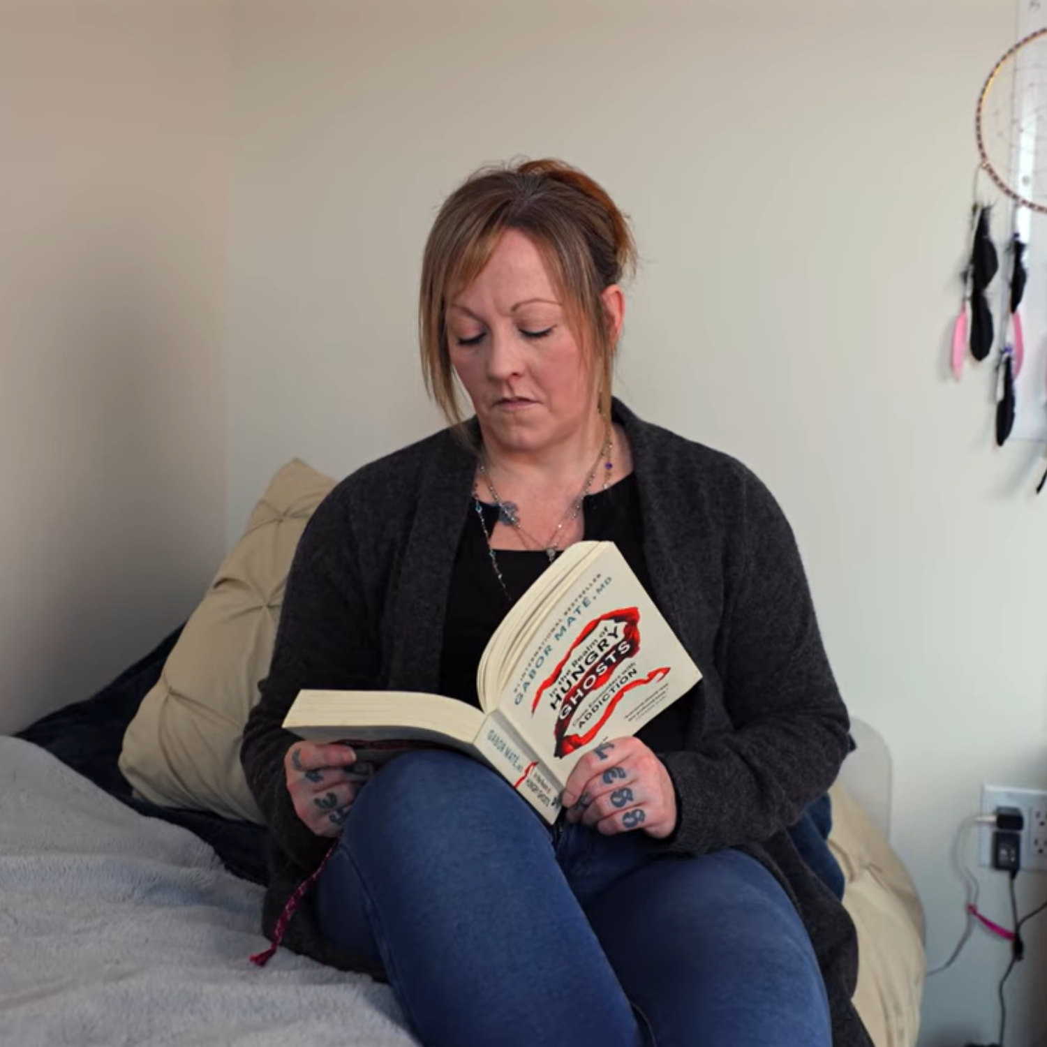 A person with blonde hair and a black shirt sits indoors on a bed while holding a white book.
