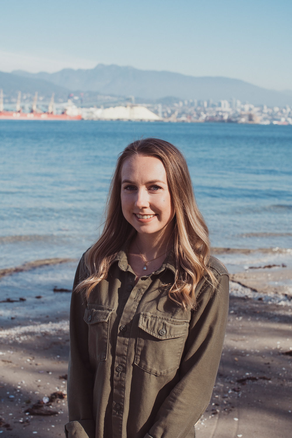 A woman with brown hair wearing a brown button up shirt poses for a picture on the beach with a body of water, a skyline and mountains in the background.