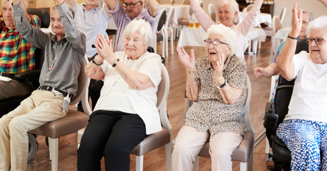 A group of cheerful seniors raise their hands while sitting down on chairs inside.