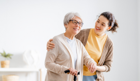 A senior using a cane is supported while walking by a younger person.