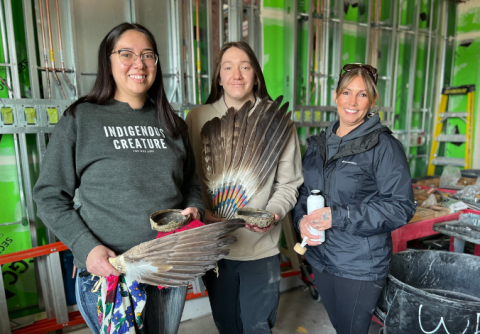 Three people dressed casually pose for a picture inside a room under construction. Two of them hold up large feathers, one of whom has a shirt reading “Indigenous Creature.”