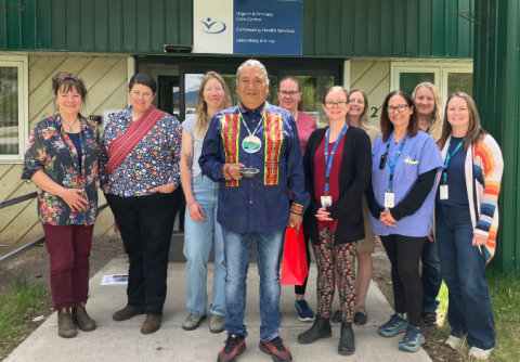 A middle-aged person dressed in some Indigenous attire holds up a couple items while posing for a group photo with other people who are dressed in medical scrubs and business casual outfits.