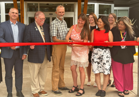 A group of happy people wearing business and business casual attire cut a large red ribbon while standing outside a building.