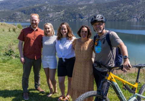 A group of people, one standing behind a bike, pose for a picture in the grass with a body of water and hills in the background.