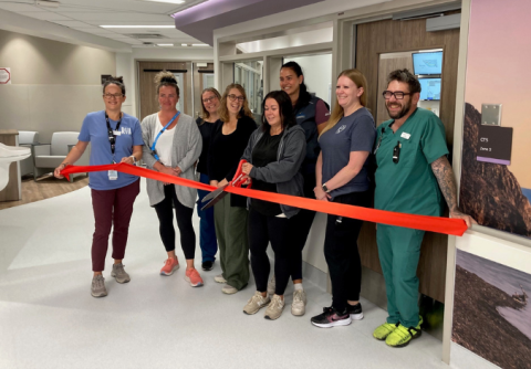 A group of happy people wearing scrubs and business casual attire cut a large red ribbon in the corner of a room.