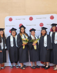 A group of six graduates in black gowns and caps and white scarves are flanked by two people in business clothing.