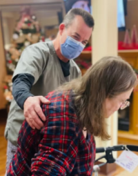 A person wearing light green medical clothes and a mask assists a person with long grey hair hunched over a walker.