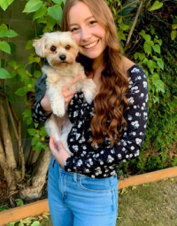 A young person with long reddish hair wearing a blouse and bluejeans holds a small cute white dog.