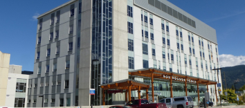 A grey six-storey hospital building with a covered breezeway of wooden pillar with three cars parked in front