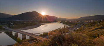 The sun rises over the mountains over two large groups of houses separated by a body of water and a bridge.