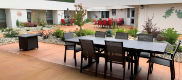 An orange patio in a courtyard with a brown set of six chairs and a table and a set of red chairs in front of a white building with flowers