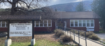An exterior view of a wide brown building with a long paved ramp leading to the front door. A side on the grass in front of the building says "Slocan Wellness Centre" and directs people to various parts of the building and lists upcoming events.