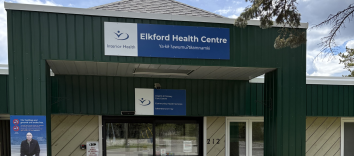 An exterior shot of a building, with an arched entrance that has a banner on it featuring the Interior Health logo and the words Elkford Health Centre.
