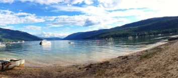 A beach and lake with boats set against a blue sky with clouds and low-lying mountains.