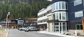 External view of a large building with vehicles parked out front. Another large building is beside it, and a tree-filled mountain range overlooks the buildings.