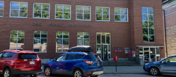 A two-storey brick building with multiple paned windows. There are several cars parked in front.
