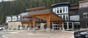 A wide building with a large wooden overhang at the front, with a forest in the background.