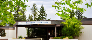 A low, one-storey beige building surrounded by trees on a cloudy day. The sign above the door says Princeton General Hospital.