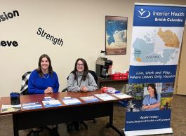 Two people sit at a table with an Interior Health promotional display next to them. The word "strength" is in the background.