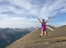 Woman wearing trail-running outfit jumping with arms overhead and a big smile, surrounded by mountains.