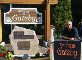 A man with grey hair leans over a platform that says “The Great Gateby” in red letters, next to a wooden sign on the left that says “Welcome to Gateby.”