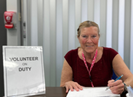 A person wearing a burgundy shirt with short sleeves sits at a table next to a volunteer on duty sign