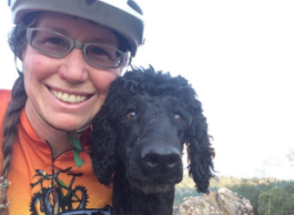 Smiling woman wearing bicycle helmet, glasses and orange t-shirt, with black poodle
