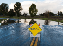 A yellow sign reading flooded sits in front of a stretch of road submerged in water.