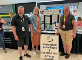 Three smiling people stand next to an Interior Health mental health and substance use sign in front of a table at an event