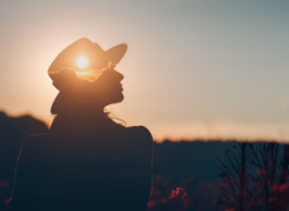 A silhouette of a person in a wide-brimmed hat sits in a field at sunset