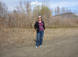A person wearing a purple shirt and black sweater and jeans stands with their hands in their pocket on a dirt road in a dry field