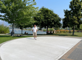 A man in a white tshirt and beige shorts stands in a large concrete semi-circle in a city park surrounded by large trees with a lake in the background
