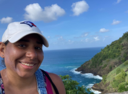 A Black woman wearing a white baseball hat, smiling with a tropical island behind her.