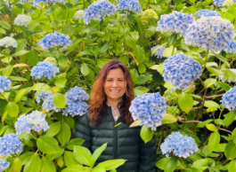 A woman with long reddish hair and green jacket stands amidst purple hydrangeas