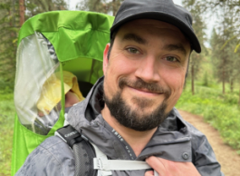 Man with dark facial hair, wearing a black baseball cap and grey rain jacket, smiles to camera. He has a child carrier on his back where a child wearing a yellow hood sleeps. He is in the forest, surrounded by trees. 