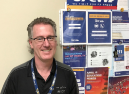 A man with curly grey hair and glasses in a black polo shirt stands next to a notice board