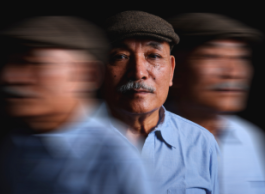 An older man with a moustache in a grey beret and wearing a blue shirt stands between two blurred images of himself