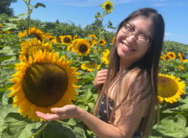 A girl with long brown hair and black tank top stands in a field of sunflowers against a blue sky