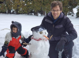 A man and child in winter gear pose for a picture outside while each resting a hand on a snowman.