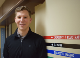 A smiling man with short sandy brown hair wearing a dark blue jacket stands in a hospital hallway with signs that say emergency, elevator and diagnostic imaging