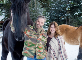 A man and woman standing in the snow with a large black horse in the snow. They are smiling for the camera and the man has one arm around the horse.