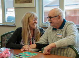 A smiling woman with long blondish hair and black sweater and plaid top grasps the hand and arm of an older man with white thinning hair, a mustache, glasses and white cableknit sweater