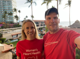 A smiling woman and man arm in arm wearing red t-shirts one which says women's heart health taking a selfie in front of a resort with palm trees and ocean in the background