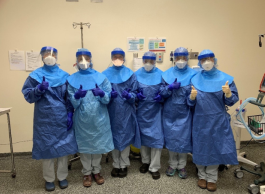 A group of six health-care professionals in blue personal protective clothing, masks and face shields all giving the thumbs up in a hospital room