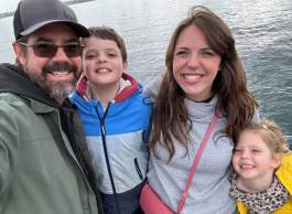 Smiling family group, man has moustache and beard, wearing sunglasses, ballcap, khaki jacket; boy wears blue and red hooded jacket with black zipper; woman has long brown hair wearing light grey pullover and pink strap shoulder bag; girl has blonde hair with white clip and yellow jacket; lake water in background
