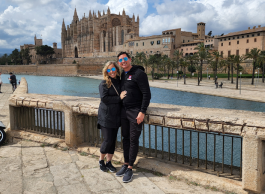 A woman and man pose for a picture on an outdoor walkway by a body of water with a castle structure in the background. 
