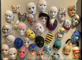 A wall of a couple of dozen brightly painted and white oval masks with two eyes, a nose and mouth