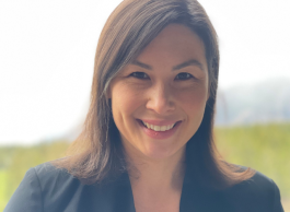 A smiling person with long straight dark hair wearing a dark blue blazer and white shirt against a backdrop of a green hill and mountain.