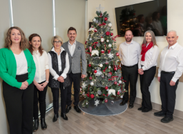 Six professionals standing in an office environment with a decorated Christmas tree at the centre.